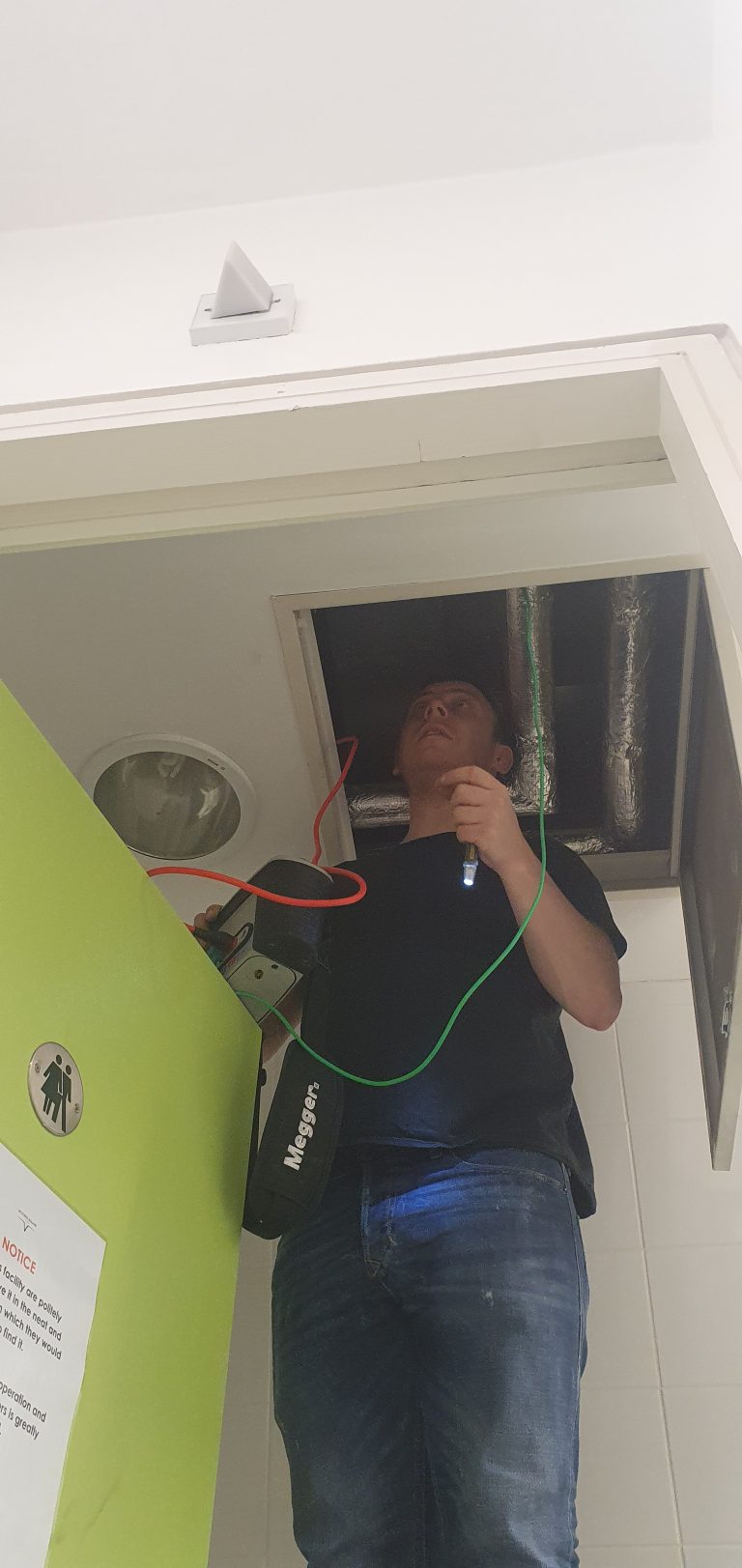 Electrician carrying out commercial electrical maintenance and inspection above a suspended ceiling in a commercial building.